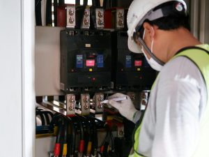 An electrical technician wearing a safety helmet and gloves inspects and marks connections inside an industrial electrical control panel with circuit breakers and heavy power cables.