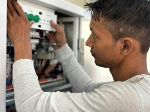 An electrician adjusts controls inside an open electrical cabinet, pressing green buttons on a control panel while inspecting internal wiring.