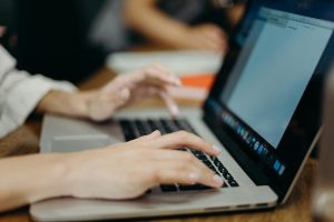 Close-up of hands typing on a laptop keyboard at a wooden table, with a document open on the screen and another person blurred in the background.