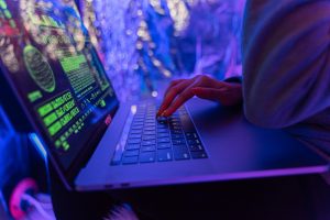 Close-up of hands typing on a laptop with neon green cybersecurity code displayed on the screen in a vibrant, purple-lit environment.