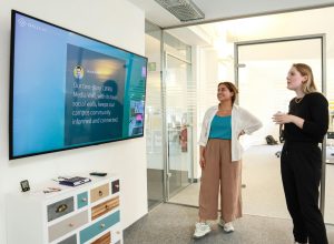 Two colleagues standing in a modern office hallway, discussing content displayed on a wall-mounted digital screen showing a social media wall, with glass meeting rooms in the background.