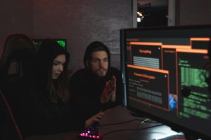 Two people in hooded sweatshirts working together in a dimly lit room, focused on computer screens displaying encryption and password-decryption interfaces.