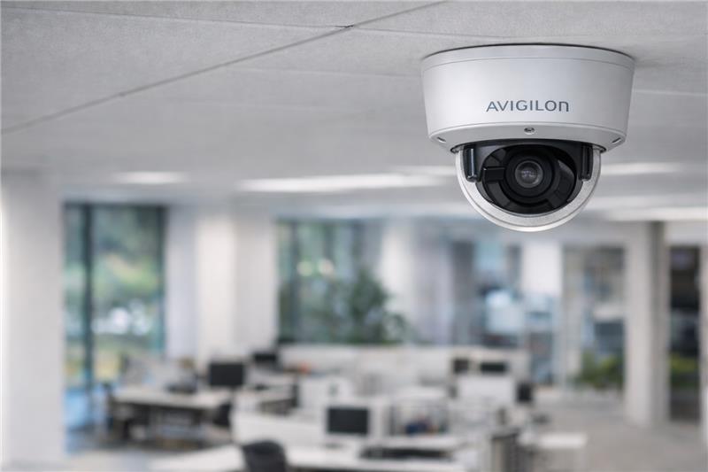Avigilon dome security camera mounted on a ceiling in a modern office environment, overlooking workstations and open-plan desks.