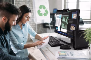 Two professionals reviewing documents while working on an Acer business desktop setup displaying analytics dashboards in a modern office environment.